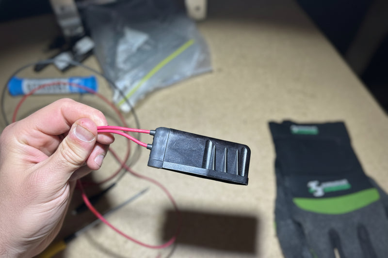 Hand holding a black electronic component with red and black wires on a textured surface.